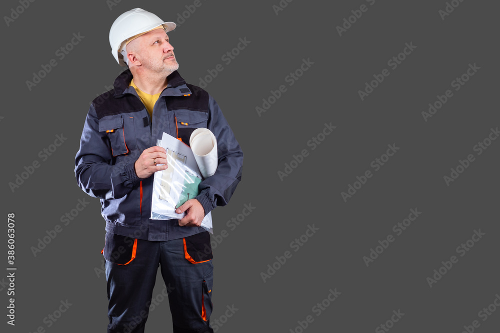 A uniformed construction worker holds drawings in his hands. Engineer in a white helmet. The architect looks up against a gray background. Architectural and construction concept with space for text.