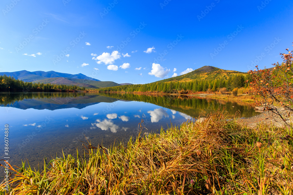 The nature of the Magadan region. A beautiful flat surface of the lake against the background of colored hills and the blue sky. Fascinating view of the forest lake