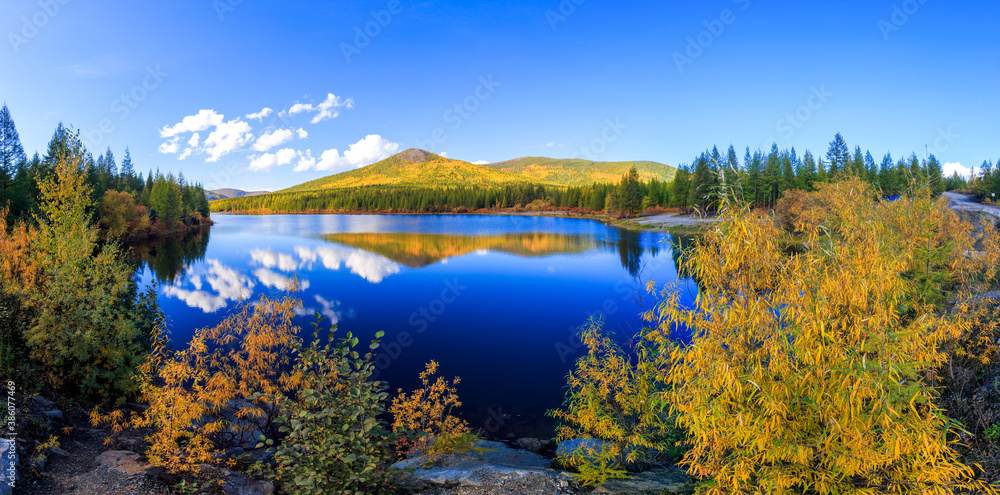 Panoramic shot. A beautiful flat surface of the lake against the background of colored hills and the blue sky. Fascinating view of the forest lake