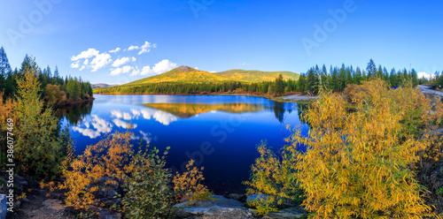 Panoramic shot. A beautiful flat surface of the lake against the background of colored hills and the blue sky. Fascinating view of the forest lake