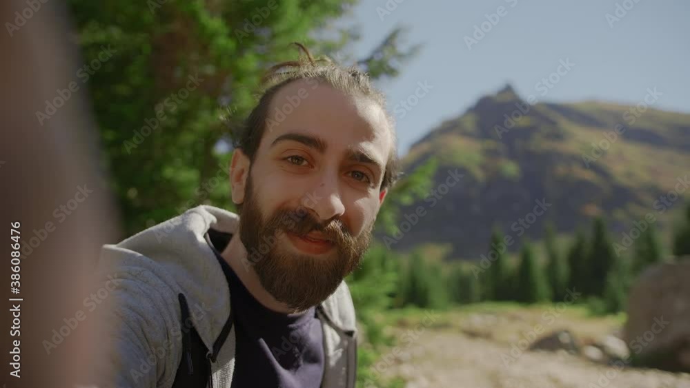 White toothed smile face hippie climber with his backpack behind him, with his handheld camera or phone, salutes the audience or the person he is talking to and points to a high mountain. White toothed smile face hippie climber with his backpack behind him, with his handheld camera or phone, salutes the audience or the person he is talking to and points to a high mountain.