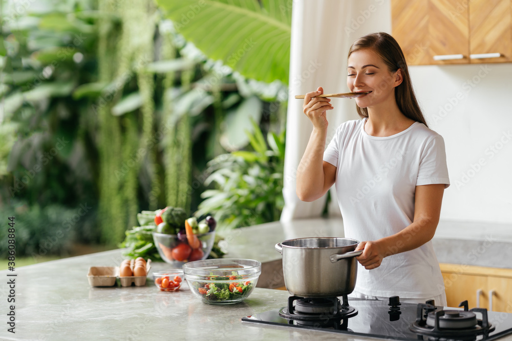 Photo of pretty caucasian woman holding cooking spoon while eating soup with fresh vegetables in open kitchen at home in Bali
