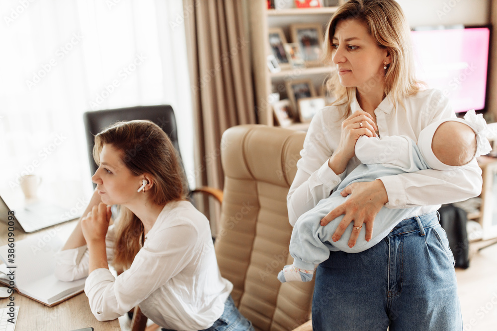 Concentrated woman sit at the desk, looking at the computer, watching webinar training, young mother working at home online, while caring grandmother babysit with newborn baby girl Concentrated woman sit at the desk, looking at the computer, watching webinar training, young mother working at home online, while caring grandmother babysit with newborn baby girl