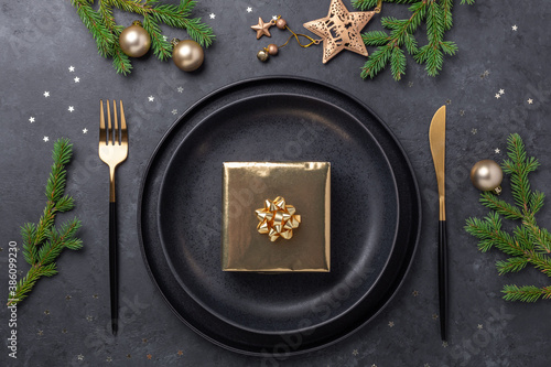 Christmas table setting. Black ceramic plate with golden gift box, fir tree branch and accessories on stone background. Gold decoration