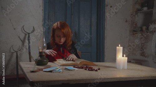 Serious pretty red haired female wizard in black cape reading ancient magic book, looking for spell while sitting at rustic table, surronded by rune stones, tarot cards and candles in dimmed house. photo