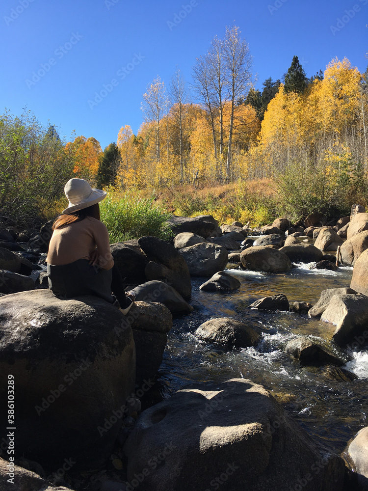 Woman in a sat sitting on a large boulder on a river, looking across at trees with fall colors, on a sunny autumn day