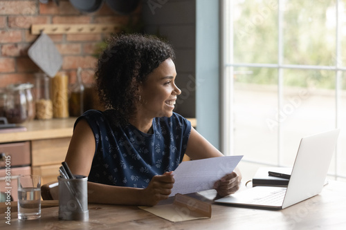 Smiling millennial biracial woman work on laptop read pleasant message in paperwork correspondence. Happy young African American female feel excited optimistic get good news in postal paper letter.