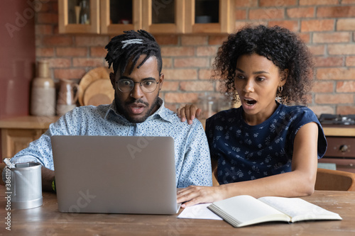 Shocked African American couple look at laptop screen frustrated by debt email paying bills online. Stunned biracial man and woman surprised by unexpected bad unpleasant news online on computer.