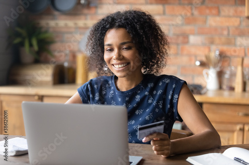 Happy young biracial woman pay online on laptop from home using bank credit card. Smiling African American female shopping on internet, make order or purchase on web with secure payment.