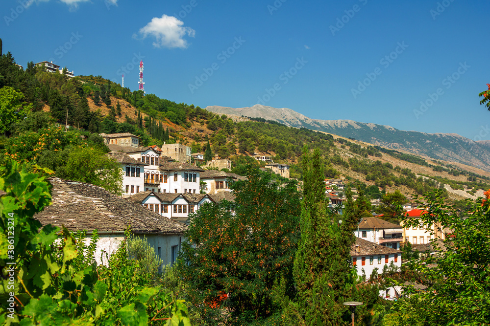 Gjirokaster town in Albania – traditional white houses with stone gray roofs on the hill. Summer landscape with lush foliage, blue sky and mountains on the horizon. 