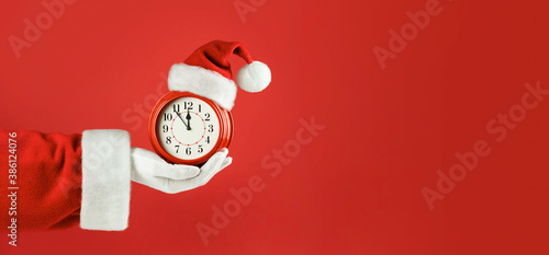Santa Claus hand holding red alarm clock in Santa hat. Red background. Concept of coming Christmas and New Year, holiday sales. Space for text, banner