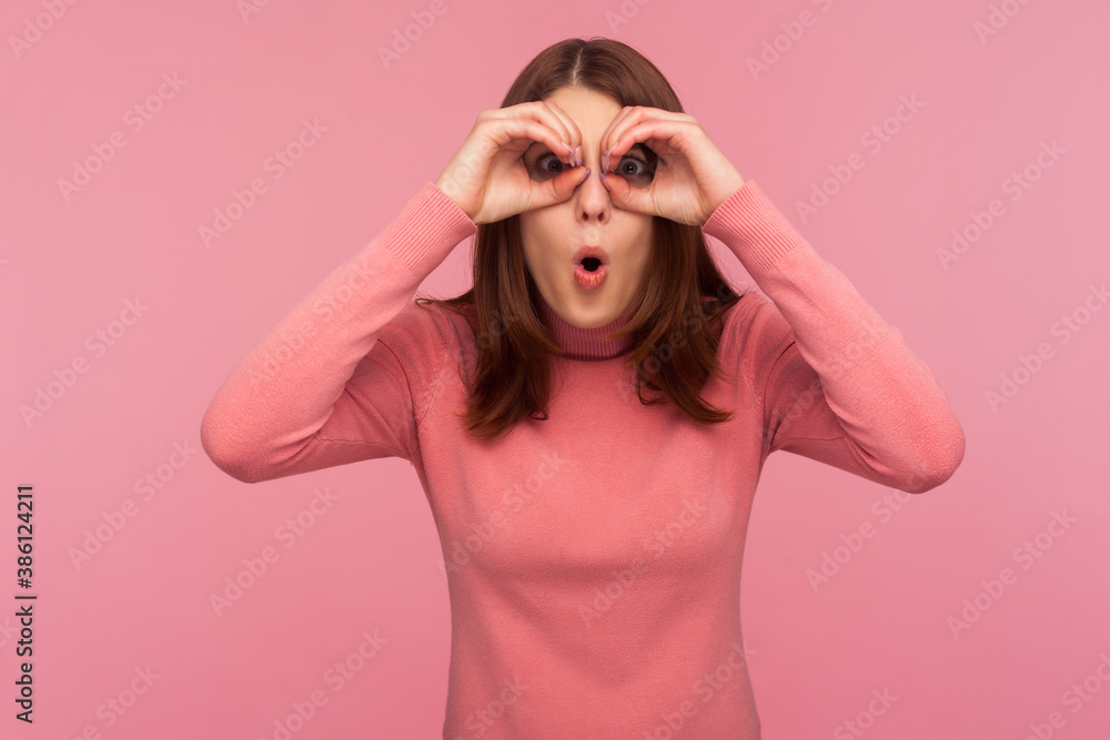 Funny curious woman with brown hair spying looking through holes in her hands, making binoculars, surprised with seen. Indoor studio shot isolated on pink background Funny curious woman with brown hair spying looking through holes in her hands, making binoculars, surprised with seen. Indoor studio shot isolated on pink background