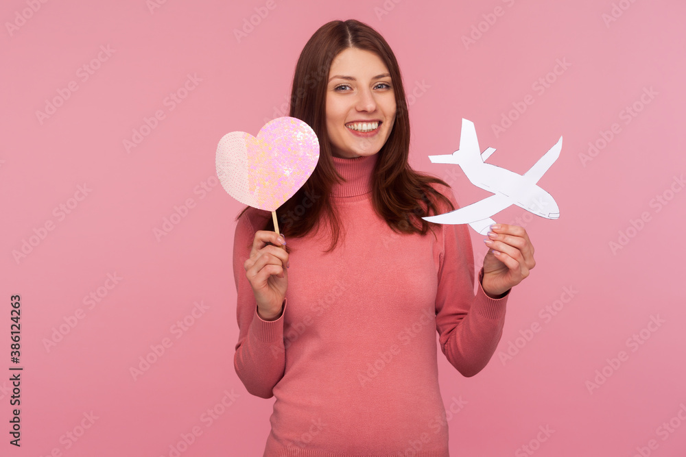 Happy cheerful woman with brown hair holding in hands paper plane and pink heart, planning honeymoon or date abroad, saint valentine day gift. Indoor studio shot isolated on pink background Happy cheerful woman with brown hair holding in hands paper plane and pink heart, planning honeymoon or date abroad, saint valentine day gift. Indoor studio shot isolated on pink background