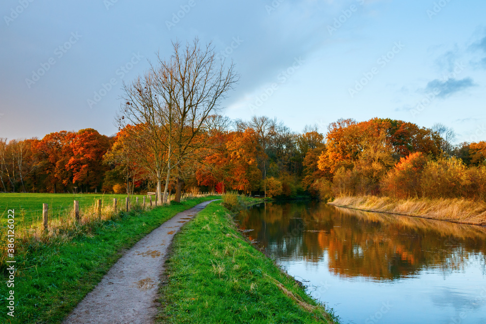 Towpath on the bank of the Kromme Rijn river (“Crooked Rhine”) with the forest of the Amelisweerd estate in autumn colors during sunset on the background.