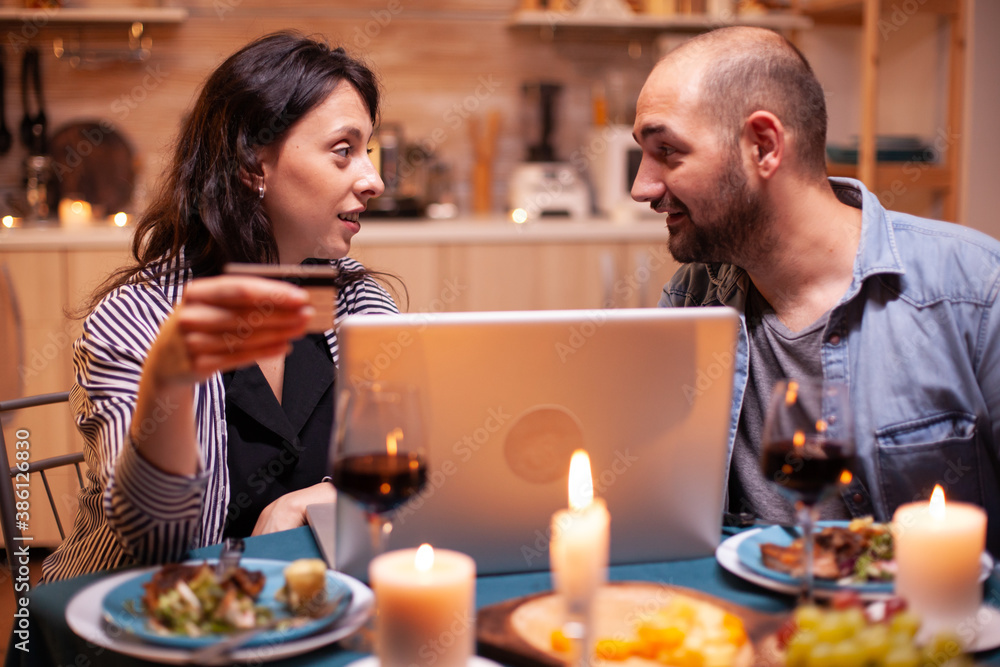Couple looking at each other while doing online shopping during romantic dinner. Adults sitting at the table, searching, browsing, surfing, using technology card payment, internet