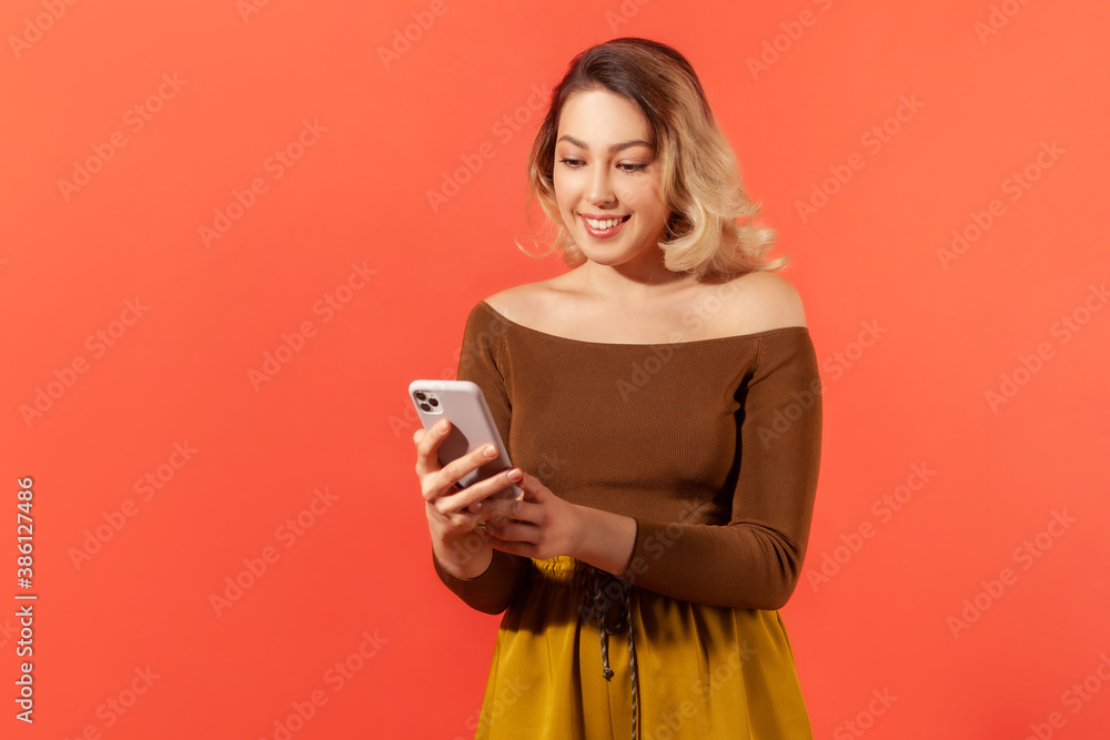 Beautiful young woman holding smartphone chatting with friends and smiling, dating app, unlimited Internet. Indoor studio shot isolated on orange background Beautiful young woman holding smartphone chatting with friends and smiling, dating app, unlimited Internet. Indoor studio shot isolated on orange background