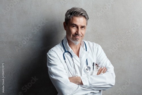 Portrait of trustworthy older confident doctor with gray hair wearing white lab coat standing against gray wall, smiling. Copy space. 