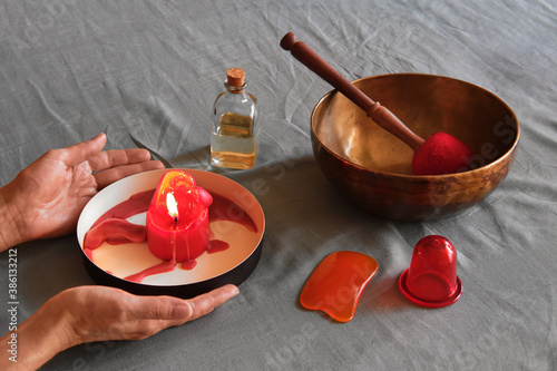 Massage items: oil, singing bowl, vacuum can on textile linen background. Female hands holding burning aroma candle