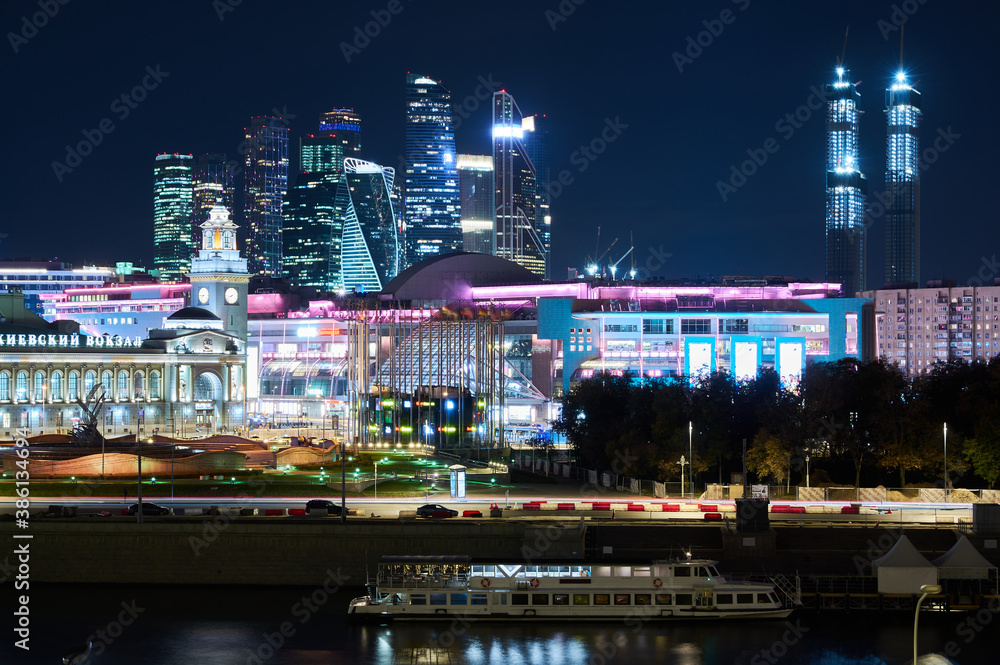 Fototapeta premium Moscow at night, sparkling with bright lights with a river and boat in the foreground and sparkling skyscrapers in the background