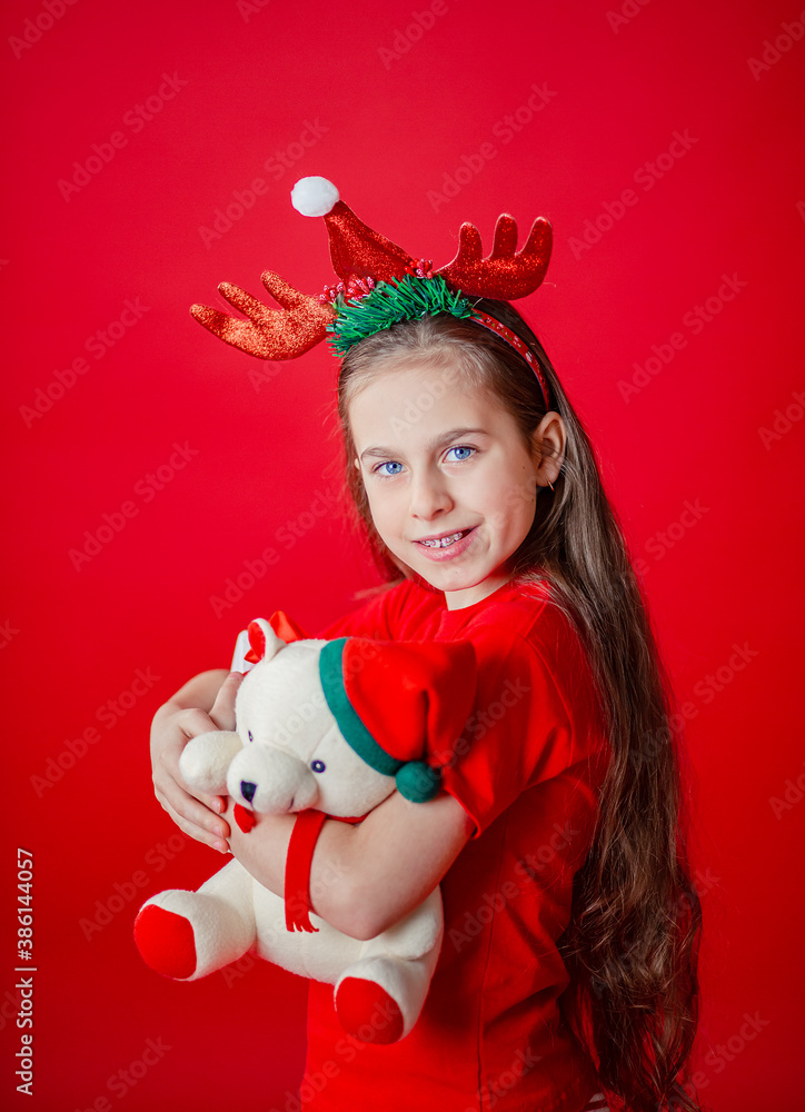 Portrait of a funny cheerful girl with a bandage of horns on her head hugging a teddy bear in Christmas pajamas isolated on a bright red background. Portrait of a funny cheerful girl with a bandage of horns on her head hugging a teddy bear in Christmas pajamas isolated on a bright red background.