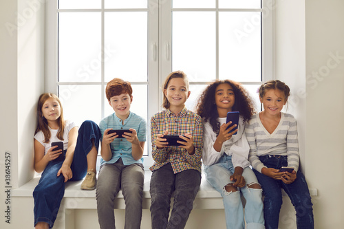 Canvas Print Happy multiethnic children playing games on mobile phones sitting on windowsill