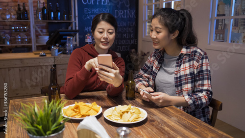 Two happy asian chinese female friends enjoying fresh bottle beer at night pub and browsing social networks on mobile phone. Technologies online communication and internet addiction concept.