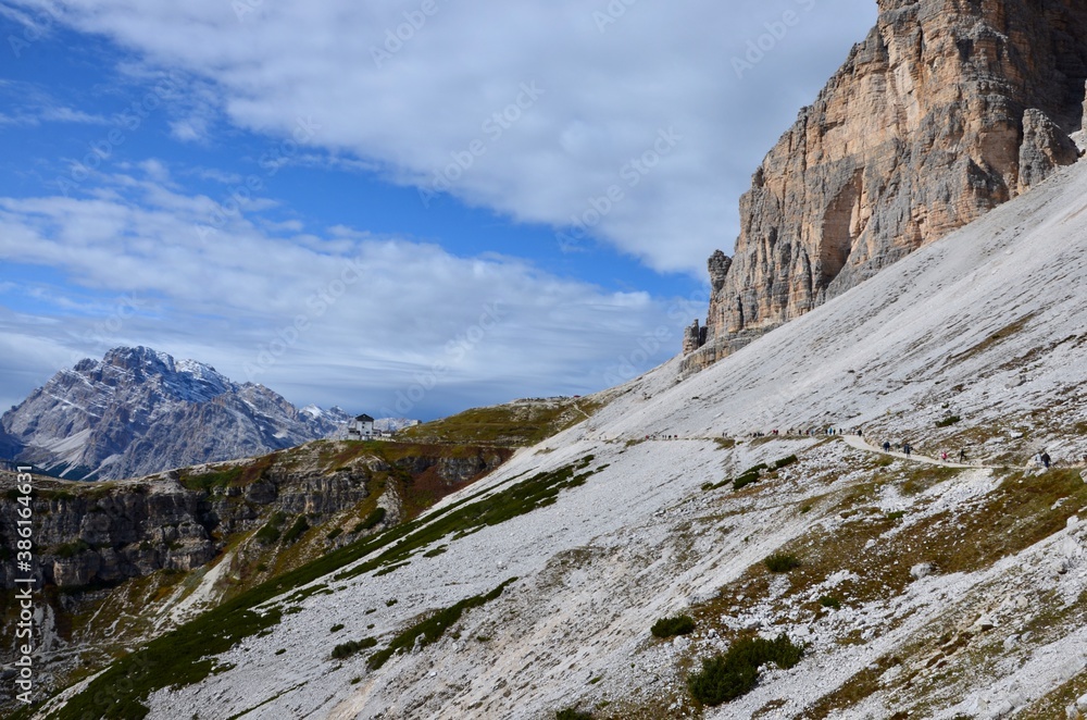 Dolomites Alps, Three Peaks (Drei Zinnen), South Tirol, Italy, UNESCO world heritage site, a footpath with hikers, mountain hut on horizon, blue sky with clouds background, a sunny day in autumn