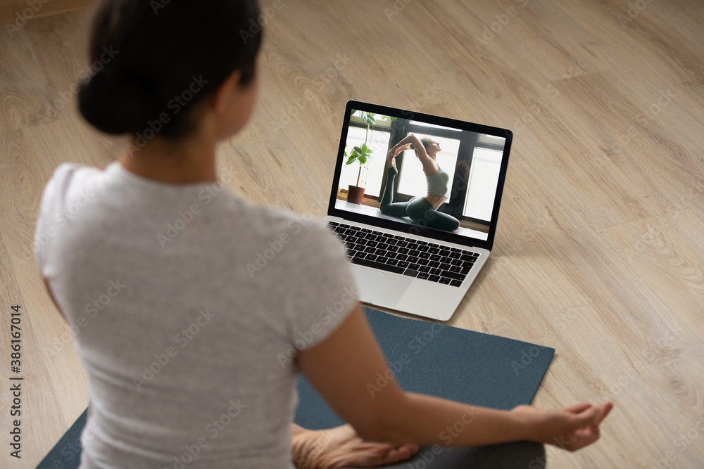 Rear view sporty Indian young woman practicing yoga at home, sitting in lotus pose on mat on floor, watching webinar online, using laptop, doing exercise, stretching, enjoying fitness activity - obrazy, fototapety, plakaty 