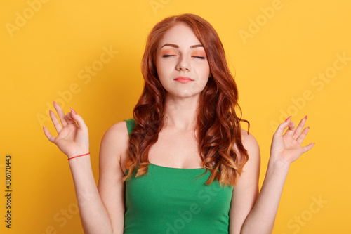Good looking woman meditates, keeps both hands in okay gesture, keeps eyes closed, relaxing after work, practices yoga, wears green shirt, isolated on green wall.
