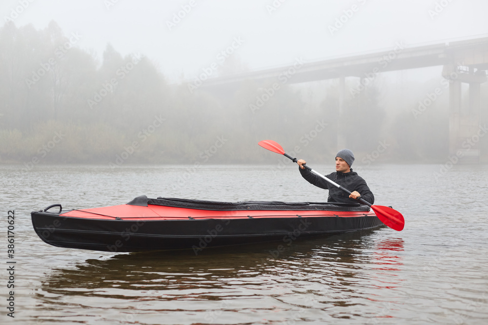 Fototapeta premium Handsome guy with oar in hands rowing his boat and looking in distance, foggy autumn morning, kayaker in middle of river with bridge on background.