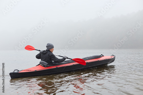 Kayaker paddling in water, holding oar in hands, sportsman in canoe, water sport, guy in jacket rowing boat in cold autumn foggy morning, active extreme rest.