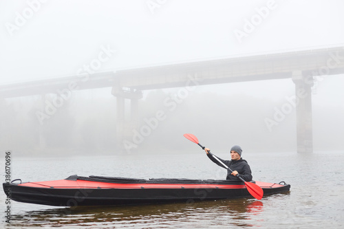 Man rowing in boat on foggy morning, holding paddle in hands, posing in water with bridge on background, looking in front of him, spending spare time in active way.