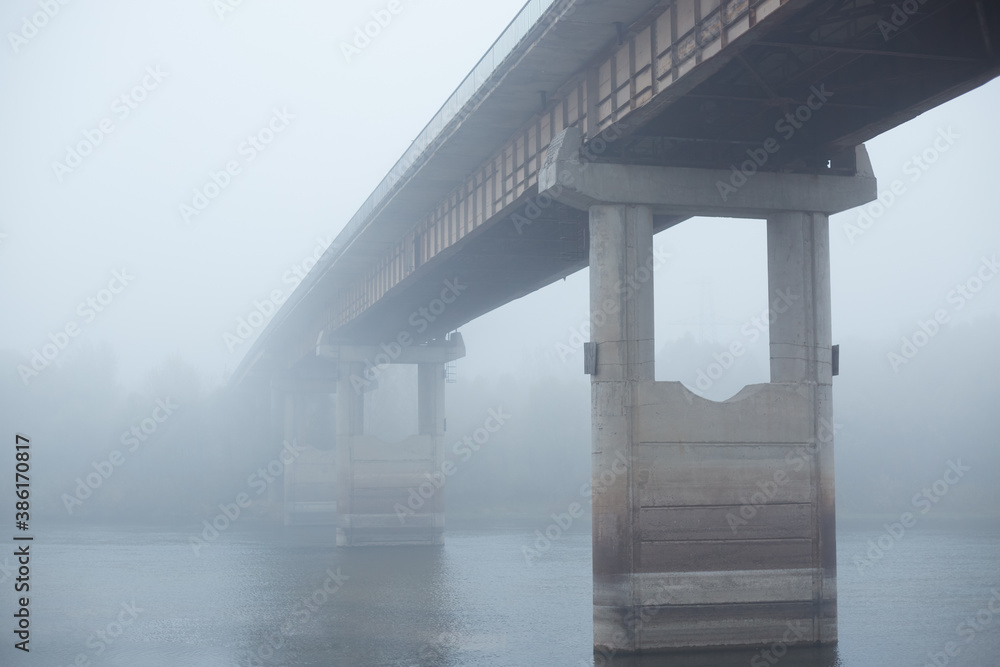 Bridge in fog, concrete bridge through river, bridgework between two banks of river, foggy sky, early morning.