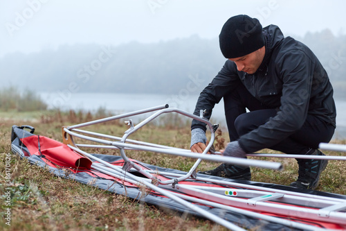 Men during kayaking, concentrated guy near canoe frame, going to padding, male in black sportswear on river bank in early foggy morning assembles kayak to travel , male near gunboat. photo