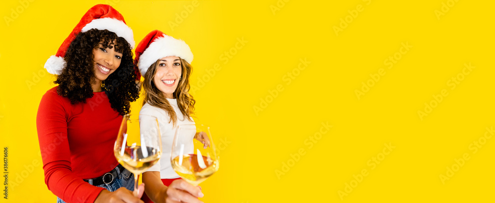 Fototapeta premium Toasting to Christmas together regardless of diversity - Two women, black Hispanic and Caucasian wearing Santa hat, looking at the camera holding a glass of white wine on big yellow copy space