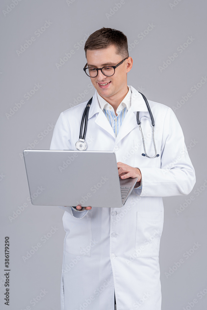 Handsome male medic is standing with laptop in hands. Smiling doctor reading good news on screen. Handsome male medic is standing with laptop in hands. Smiling doctor reading good news on screen.