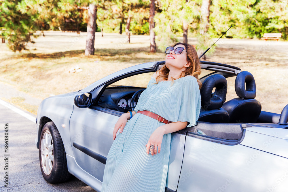 Outdoor fashion portrait. Attractive woman leaning and posing at convertible car. Happy pretty girl. Relaxing on summer day in nature