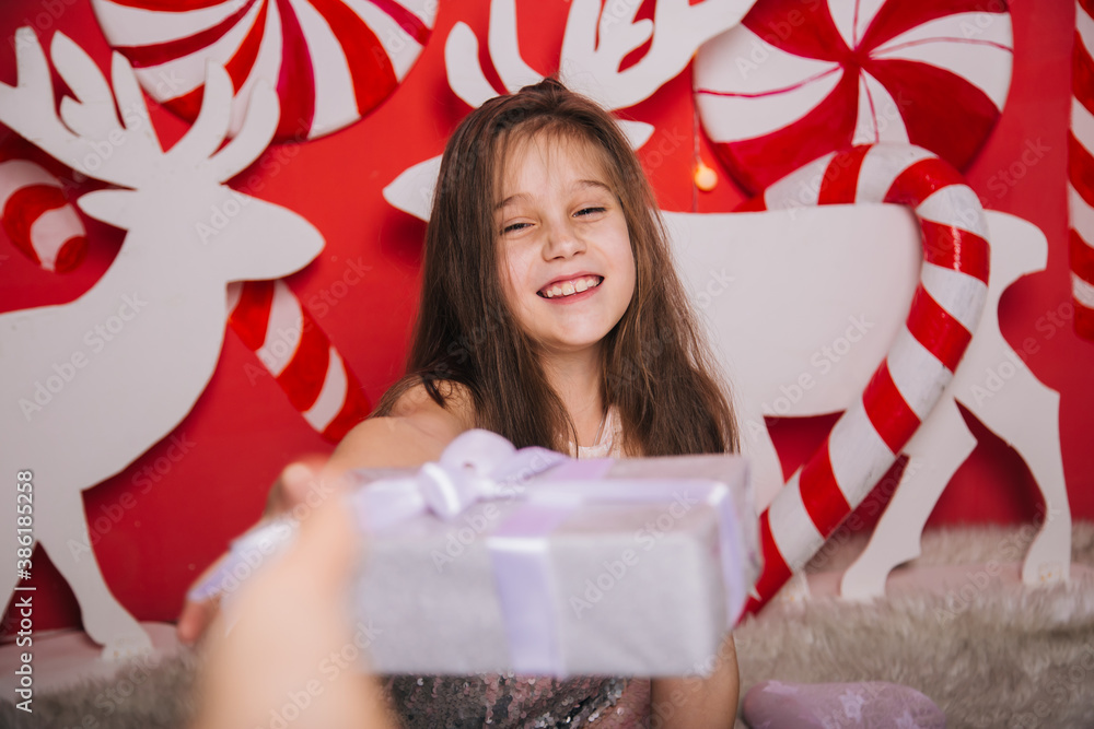 Cheerful girl holding a Christmas present in her hands. A wall of lollipops. A child sits on a carpet against the background of New Year's decorations with deer Cheerful girl holding a Christmas present in her hands. A wall of lollipops. A child sits on a carpet against the background of New Year's decorations with deer