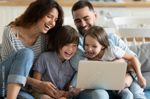 Overjoyed parents with little son and daughter using laptop together, looking at screen, sitting on cozy couch at home, happy mother and father with two kids making video call, having fun with device