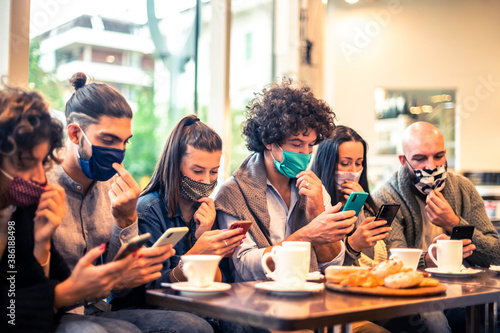 Multiracial friends with face mask holding smartphones sitting at table drinking cappuccino at cafe shop in the covid-19 time
