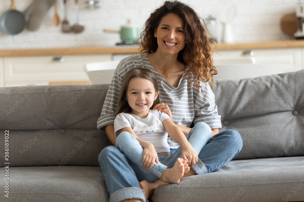 Obraz premium Portrait smiling young mother hugging little daughter, looking at camera, positive happy beautiful woman mum and cute adorable girl kid sitting on couch at home, posing for photo together