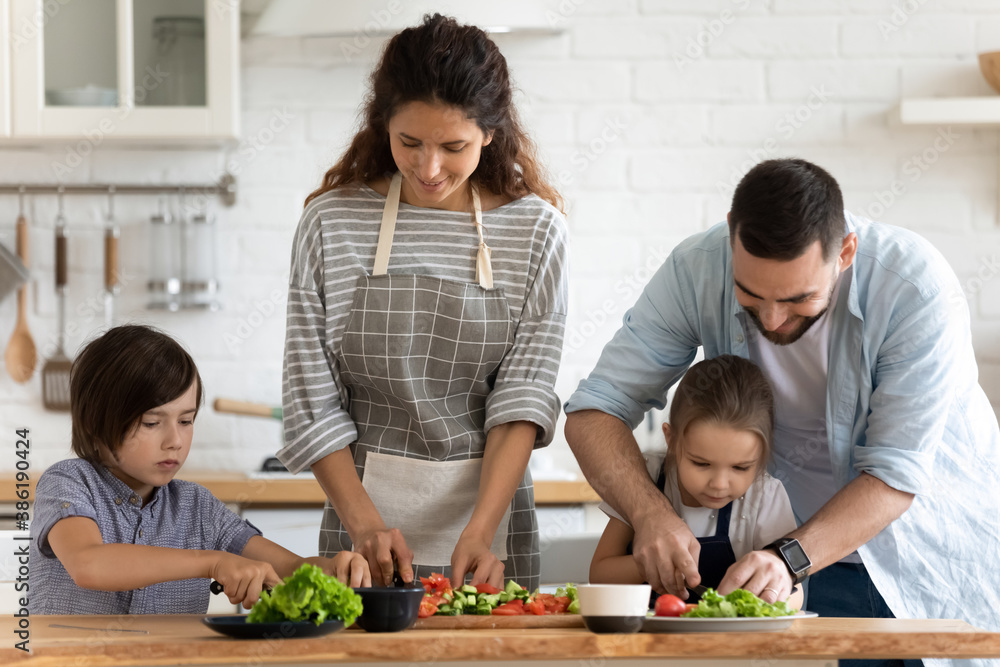 Happy young family with two adorable little kids cooking salad together, caring father teaching daughter to cut fresh vegetables, parents with children preparing dinner in modern kitchen