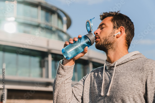 Low angle view of a handsome 20s male jogger drinking water after running against urban style building