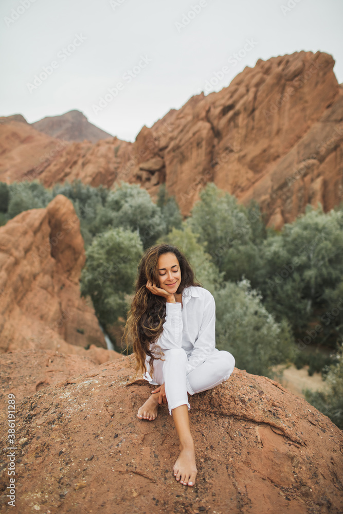 Pretty brunette woman in white sitting and enjoying view of Todra gorge canyon in Morocco. Harmony with nature and freedom concept. Travel lifestyle.
