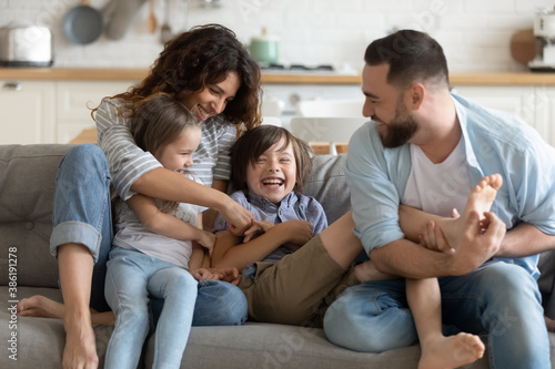 Happy young parents with two kids having fun at home, laughing overjoyed mother and father tickling little daughter and son, sitting on couch in living room, family enjoying leisure time together