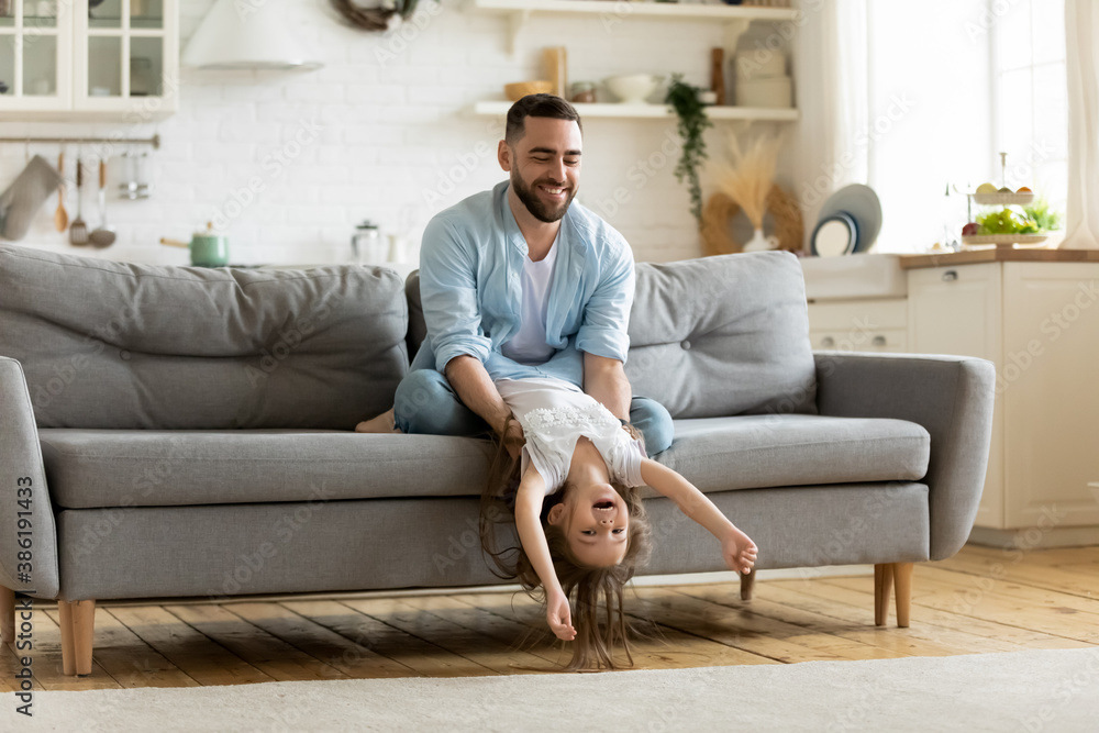 Loving young father playing with daughter at home, happy dad holding cute little girl upside down, sitting on cozy couch in living room, family playing funny game, spending leisure time together Loving young father playing with daughter at home, happy dad holding cute little girl upside down, sitting on cozy couch in living room, family playing funny game, spending leisure time together