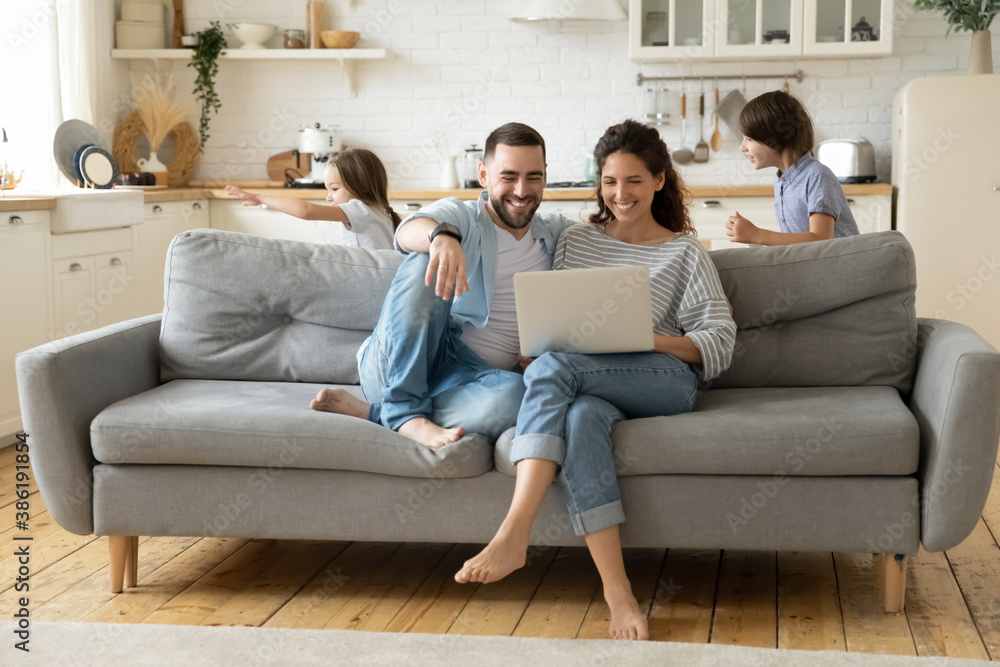 Happy parents using laptop, sitting on couch while kids playing funny game, smiling mother and father shopping online, enjoying leisure time, little son and daughter running and laughing together Happy parents using laptop, sitting on couch while kids playing funny game, smiling mother and father shopping online, enjoying leisure time, little son and daughter running and laughing together