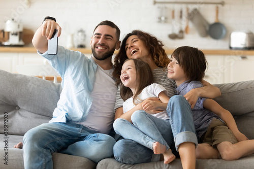 Happy parents with adorable little kids having fun, using mobile device, smiling father holding smartphone, taking selfie, overjoyed young family with daughter and son sitting on cozy couch at home
