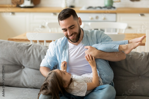 Happy young father playing with adorable little daughter at home, sitting on cozy couch at home, loving dad holding cute girl, hugging and tickling, having fun, family enjoying leisure time together