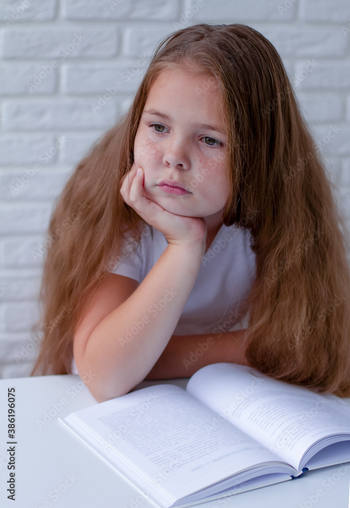 An angry and tired schoolgirl studies with a book on her Desk. Unwillingness to learn. Look out the window An angry and tired schoolgirl studies with a book on her Desk. Unwillingness to learn. Look out the window